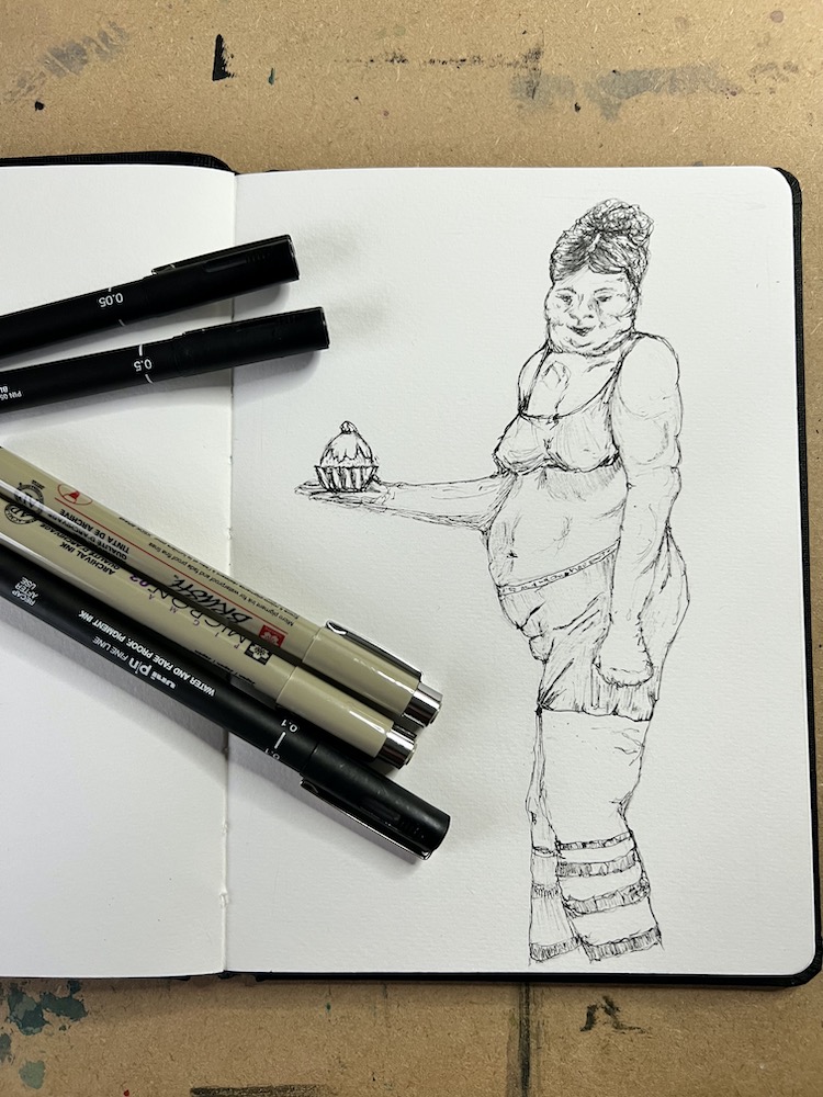 sketch book and pens with a drawing in ink of a fat lady with a cake